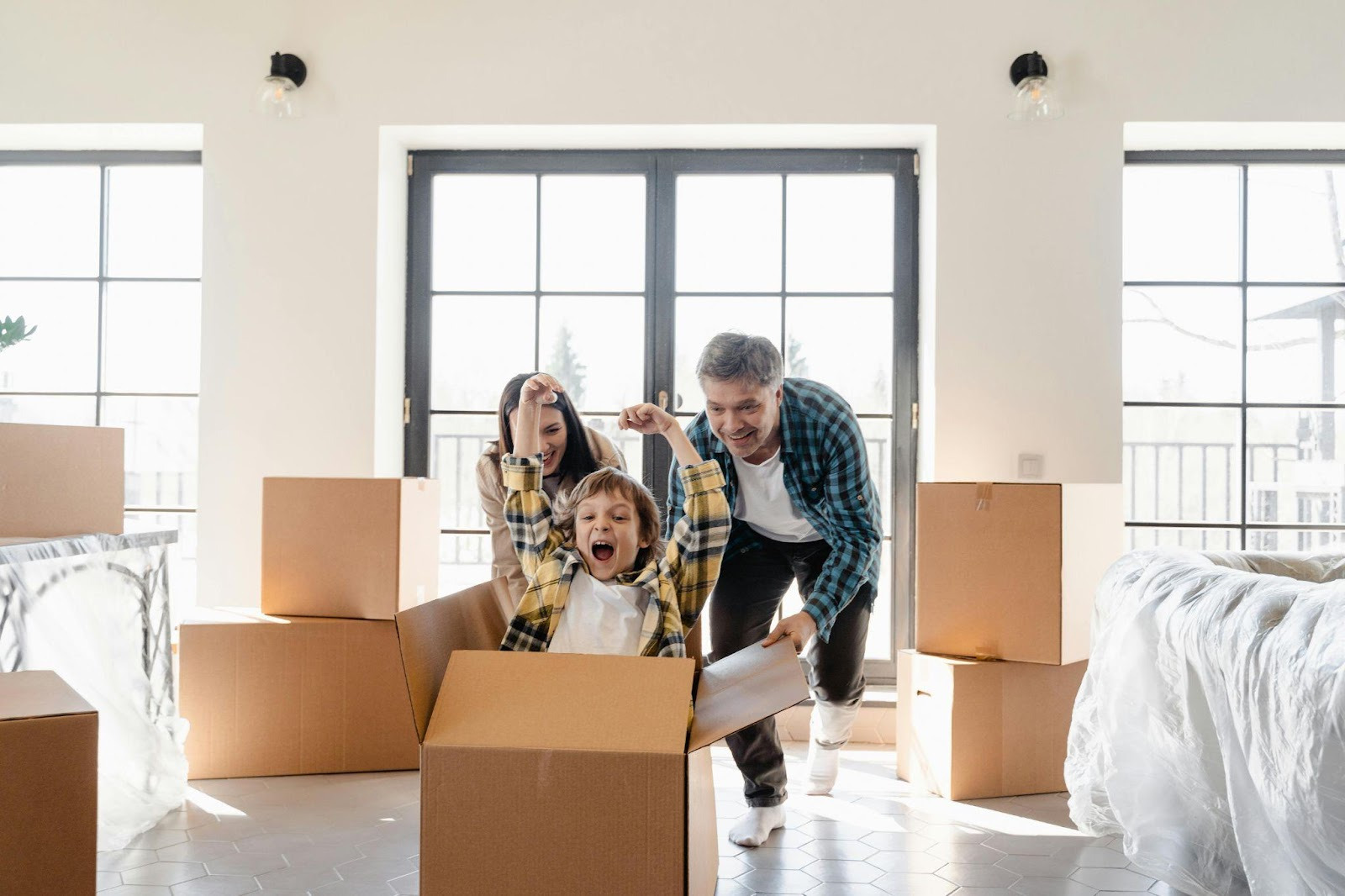 A joyful family, father, mother, and child, playfully interacts with moving boxes in a bright room. The child laughs from inside a box, conveying excitement.